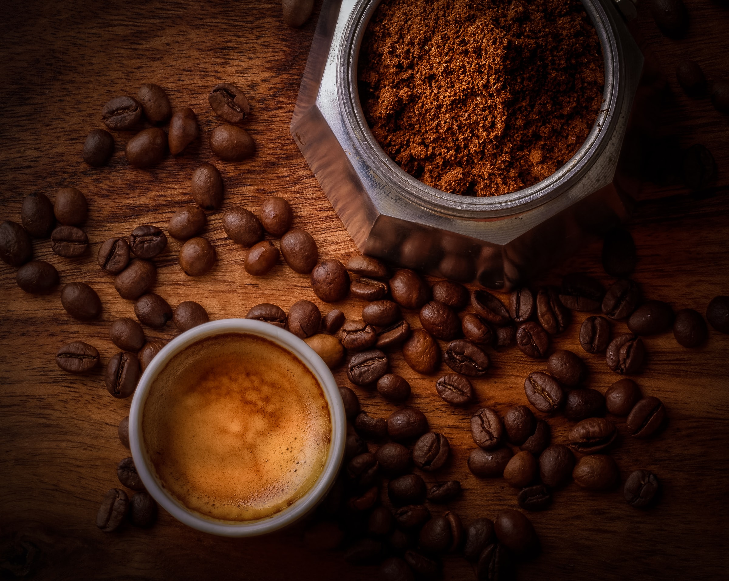 Cup of espresso next to an espresso pot filled with ground coffee, surrounded by whole roasted coffee beans.